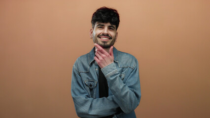 Fototapeta premium Young man with a denim jacket smiles confidently in front of a plain brown background, exuding charm and friendliness.