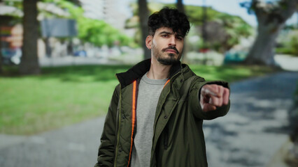 Young man with beard wearing jacket pointing in a park setting with trees and buildings blurred in...