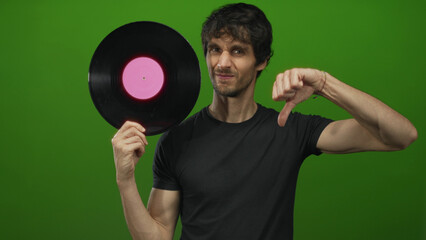 Man holds vinyl record with pink label and shows a thumbs down gesture in a vibrant green studio;...