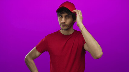 Man in red shirt adjusts red cap with one hand and places the other on hip in studio with magenta...