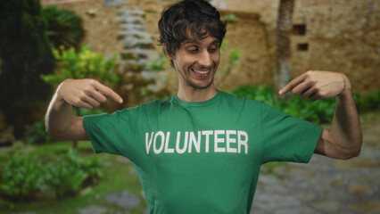Man smiling and finger pointing to volunteer shirt in green sunlit park outdoors near a stone...