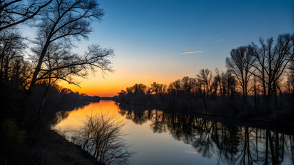 Obraz premium A tranquil river at sunset, framed by trees, with vibrant hues reflecting on the calm water