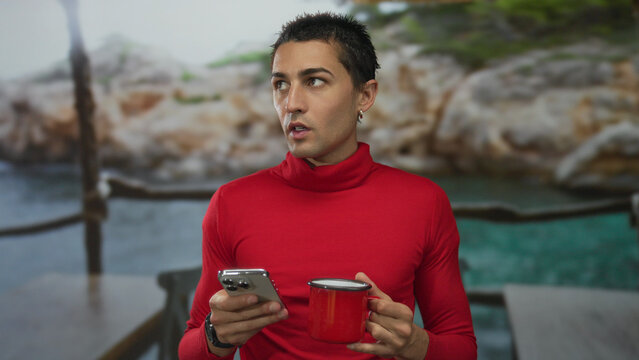 Young hispanic man holding smartphone and coffee cup at an outdoor restaurant terrace with a scenic background.