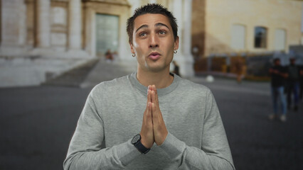 Young man in old town outdoors, gesturing with hands in prayer near historical architecture,...