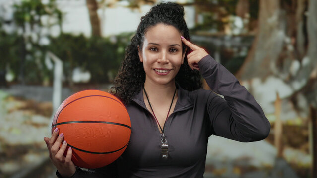 Woman holding basketball outdoors touches head playfully, suggesting an idea in a sunny park environment, displaying hispanic heritage and wearing sports attire with a whistle. - Powered by Adobe