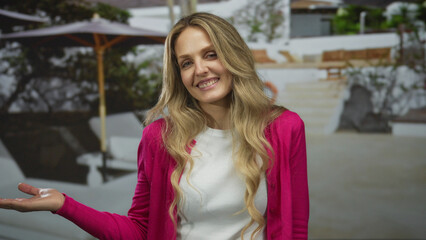 Blonde woman smiling outdoors in a garden setting with umbrella providing shade, wearing a vibrant pink jacket and white shirt, embodying joy and relaxation in natural surroundings.