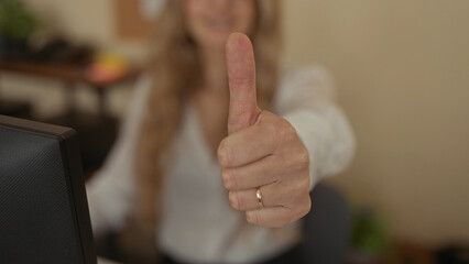 Woman gesturing thumbs in office setting giving approval and disapproval with hand showing young blonde female in workplace environment indoors interior room computer desk.