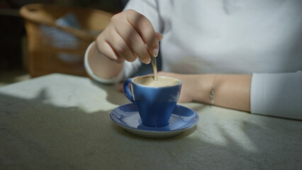 Woman stirring coffee in a blue cup at a mediterranean restaurant, showcasing a relaxed indoor setting bathed in natural light and shadows on a stone table.
