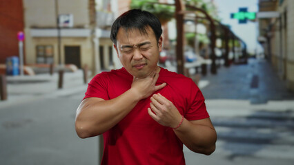 Man with pained expression touches his throat while standing outside on a city street wearing a red...