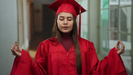 Young woman in red graduation gown meditating in hotel hallway, symbolizing calm and achievement...