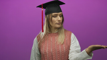 Young woman with blonde hair wearing graduation cap gestures no with finger against isolated purple...