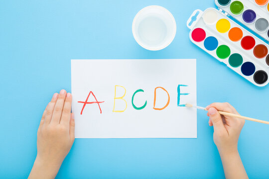 Little child holding paintbrush and painting colorful different english letters on white paper with watercolor on blue table background. Pastel color. Closeup. Creativity learning. Top down view.