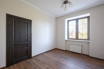 Empty room with hardwood floors, a brown door, and a window letting in natural light. Walls are light beige