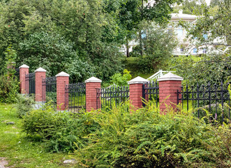 A wrought metal black fence with lattice and red brick posts separates the park from the lady on the hill behind the trees