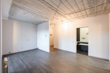 An empty room with gray floors, white walls, exposed concrete ceiling, and a bathroom visible through a doorway