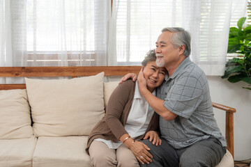 Fototapeta premium Asian couple elderly embracing together while sitting on cozy sofa at home. Happy senior retirement hug in love.