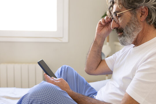 Side view of mature man using phone relaxing on bed at home.
