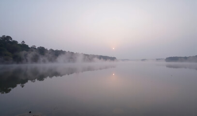 A misty sunrise over a lake with trees in the background.