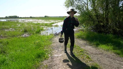 A fisherman walks up a green grassy hill to a lake with a fishing rod and a bucket, wearing a cowboy hat, a plaid shirt, grey trousers and rubber boots. It is a sunny day outside and there are a lot o