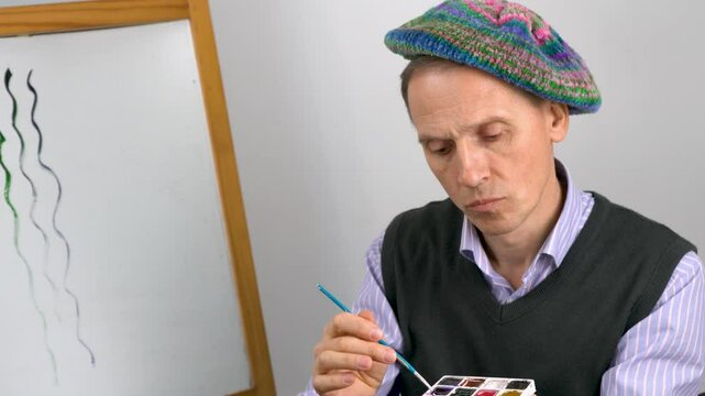 An elderly male artist in a striped blue shirt, a gray sweater and a knitted colored beret carefully stirs paint with a brush.