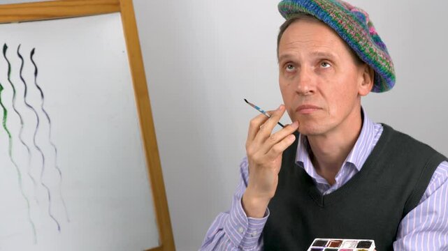 An elderly artist in a striped blue shirt, a gray sweater and a knitted colorful beret is thinking about painting a picture.