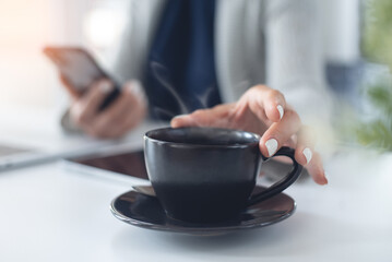 Taking a coffee break. Business woman using mobile phone during working on laptop computer, hand holding a cup of coffee on office table, closeup