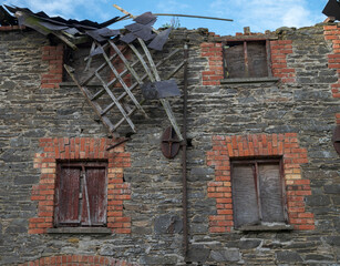 The wall of an abandoned industrial building shows crumbling stone and brick, with boarded-up windows and ivy climbing the surface. Photo taken in Inniskeen, Ireland