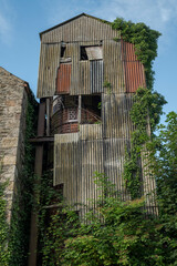 Ruined tower covered with corrugated asbestos roofing sheets and overgrown with ivy. Its weathered, crumbling walls reflect a state of neglect and industrial decay. Photo taken in Inniskeen, Ireland