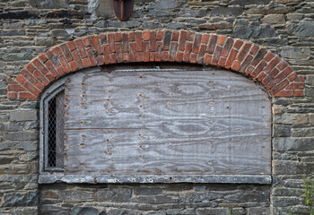 The wall of an abandoned, industrial building shows crumbling stone and brick, with boarded-up windows, evoking a sense of decay and neglect. Photo taken in Inniskeen, County Monaghan, Ireland