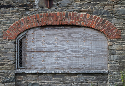 The wall of an abandoned, industrial building shows crumbling stone and brick, with boarded-up windows, evoking a sense of decay and neglect. Photo taken in Inniskeen, County Monaghan, Ireland
