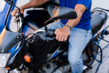 A close-up shot of a Southeast Asian tricycle driver’s hand gripping the motorcycle handlebar, wearing a blue shirt and jeans, ready to drive.