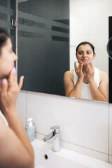 Young woman applying moisturizing cream to her face in the bathroom in front of a mirror, daily...