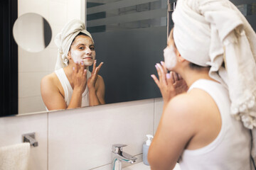 Young woman cleansing and scrubbing her face with cosmetics in the bathroom in front of a mirror, facial skincare routine