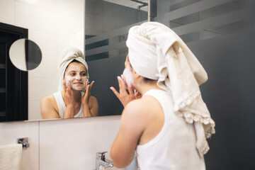 Young woman cleansing and scrubbing her face with cosmetics in the bathroom in front of a mirror, facial skincare routine