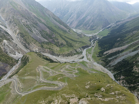 Aerial view of a winding road snaking through a lush, green valley framed by steep, rocky mountains under a cloudy sky, Kirpichny, Issyk-Kul Region, Kyrgyzstan.