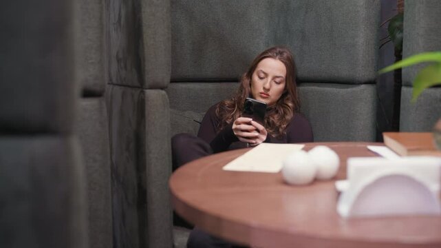 Young woman dressed in black sits on cushioned booth with legs crossed while operating phone, wooden table with decorative items placed beside her, relaxed indoor setting with calm focused expression
