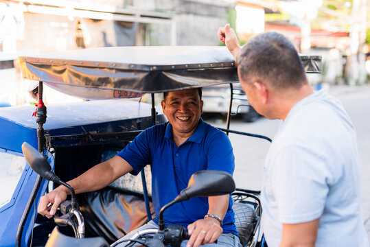 Middle-aged Southeast Asian tricycle driver in a blue shirt smiling warmly as he engages in cheerful conversation with a customer by the roadside in a sunny local neighborhood.