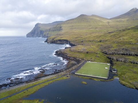 Aerial view of a football field nestled between the rugged coastline and a serene lake, a striking contrast of green against the deep blues, Funningsfj&Atilde;&cedil;r&Atilde;&deg;ur, Eysturoy, Faroe Islands.