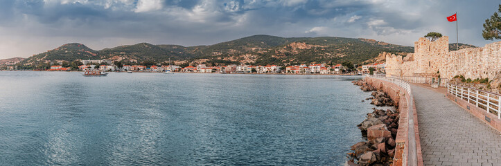 Coastal pathway beside calm waters, leading to a historic fortress in Foca with a Turkish flag,...