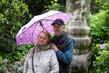 Elderly couple standing under an umbrella in a summer park. Concept: enduring love, togetherness,...