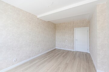 empty room with patterned beige wallpaper, light wood floors, and a white door. The ceiling is white with a recessed section. Outlets are visible along the walls
