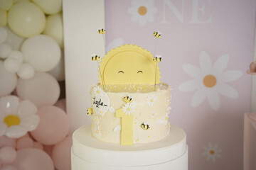 Charming first birthday cake with a smiling sun, fondant bees, and daisies, styled with soft pastel...