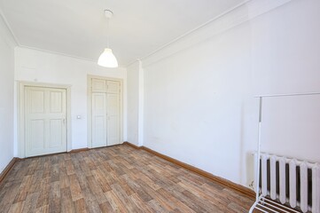 bright, empty room with white walls and wood-look flooring. Two doors are on the left, a radiator and clothing rack on the right, and a simple pendant lamp hangs from the ceiling