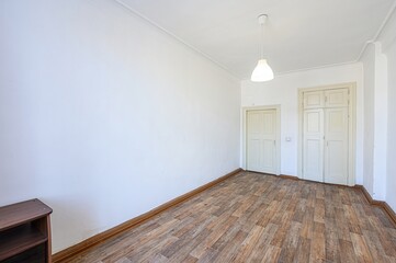 empty room with wooden floors and white walls. There are two white doors and a dark wooden shelf in the corner. A simple light fixture hangs from the ceiling