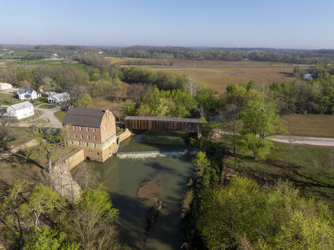 Aerial view of the sunlit mill building casting shadows on the flowing river, with a covered bridge standing guard, Missouri, United States.