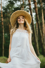 Smiling woman wearing white dress and straw hat walking in forest