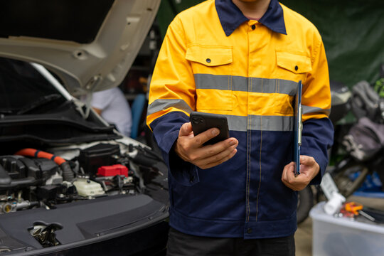Mechanic in yellow jacket using smartphone near open car hood