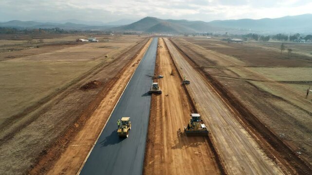 A wide construction zone with machines building a road across open land, captured from above to show planning, development, and heavy infrastructure equipment