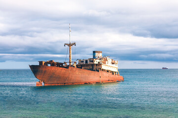 Rusty abandoned ship stranded in calm turquoise sea under dramatic cloudy sky, evoking a sense of...