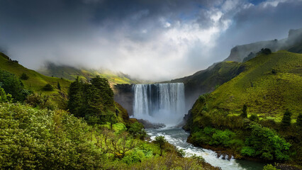 Fototapeta premium Iconic Waterfall with Rainbow Spray Falling from Sheer Cliff Surrounded by Yellow Wildflowers and Blue Sky, Perfect for Nature Tourism and Travel Photography Backgrounds